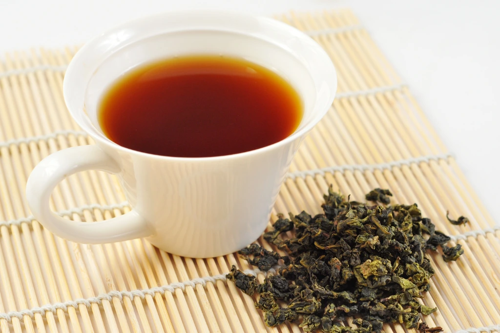 A cup of tea with dried tea leaves at the side on a wooden mat