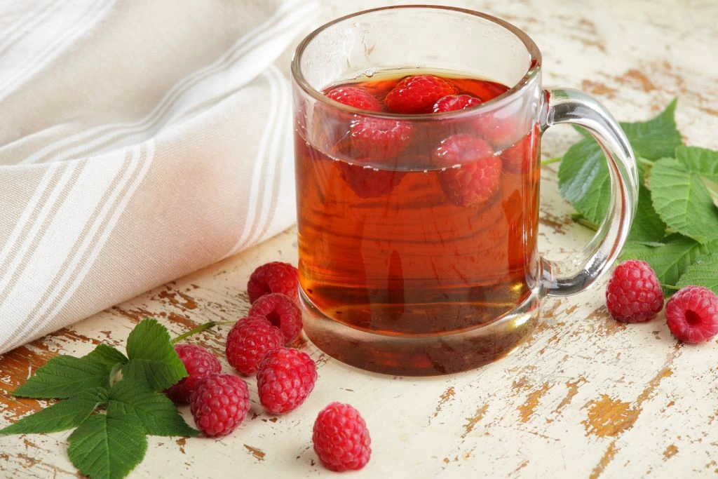 Glass cup filled with red raspberry tea surrounded by red raspberry fruit