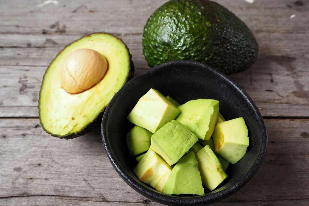 Two avocado fruits and a bowl of sliced avocado on a table