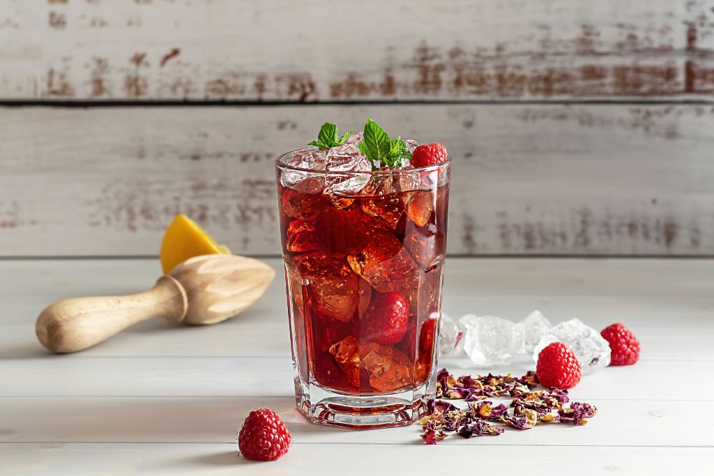 a glass of hibiscus iced tea with raspberries, mint, and lemon on a wooden table