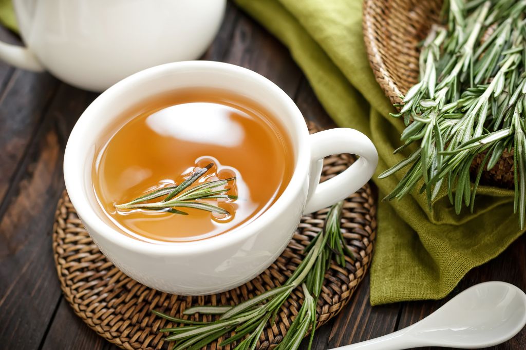 a white cup with rosemary tea together with fresh rosemary leaves beside it