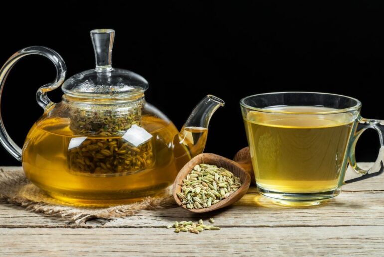 Fennel tea in glass cup and a glass tea pot with dried fennel seeds along with a wooden spoon with fennel seeds in it